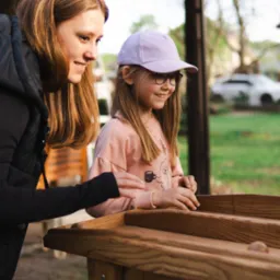 Dziewczynka w fioletowej czapce i okularach gra w drewnianą grę z kulkami, uśmiechając się, obok stoi kobieta w czarnej kurtce, obserwując grę.
