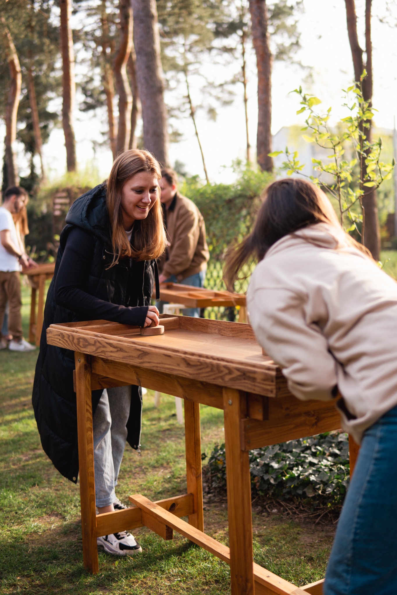 Gra plenerowa: Uśmiechnięta kobieta w czarnej kamizelce gra w drewnianą grę z krążkami na zewnątrz, w tle widoczni inni gracze i drzewa.