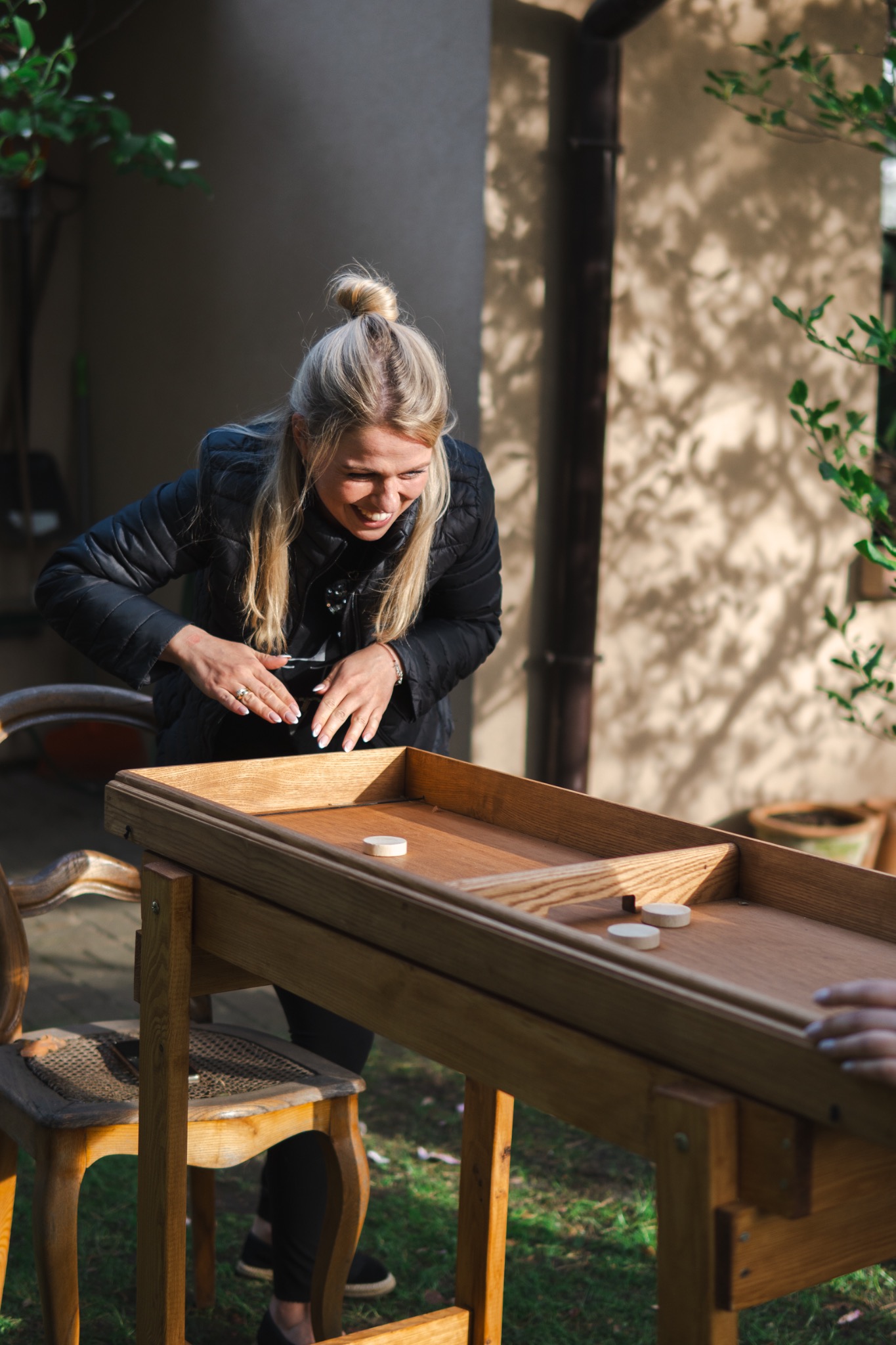 Uśmiechnięta blondynka w kurtce gra w drewnianą grę z krążkami na zewnątrz. Widoczne ręce drugiej osoby i elementy otoczenia.