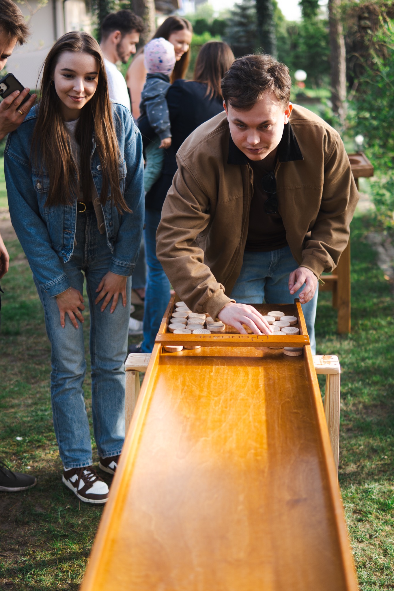 Gra w plenerze: Mężczyzna ustawia drewniane krążki na pochylonej planszy. W tle obserwują uśmiechnięci ludzie, w tym kobieta w jeansowej kurtce i dziecko na rękach.