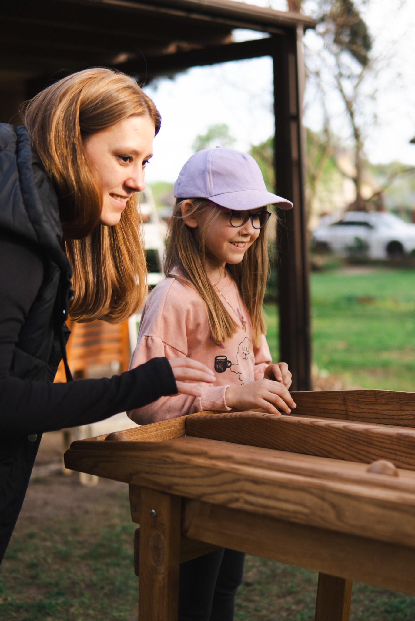 Dziewczynka w fioletowej czapce i okularach gra w drewnianą grę z kulkami, uśmiechając się, obok stoi kobieta w czarnej kurtce, obserwując grę.