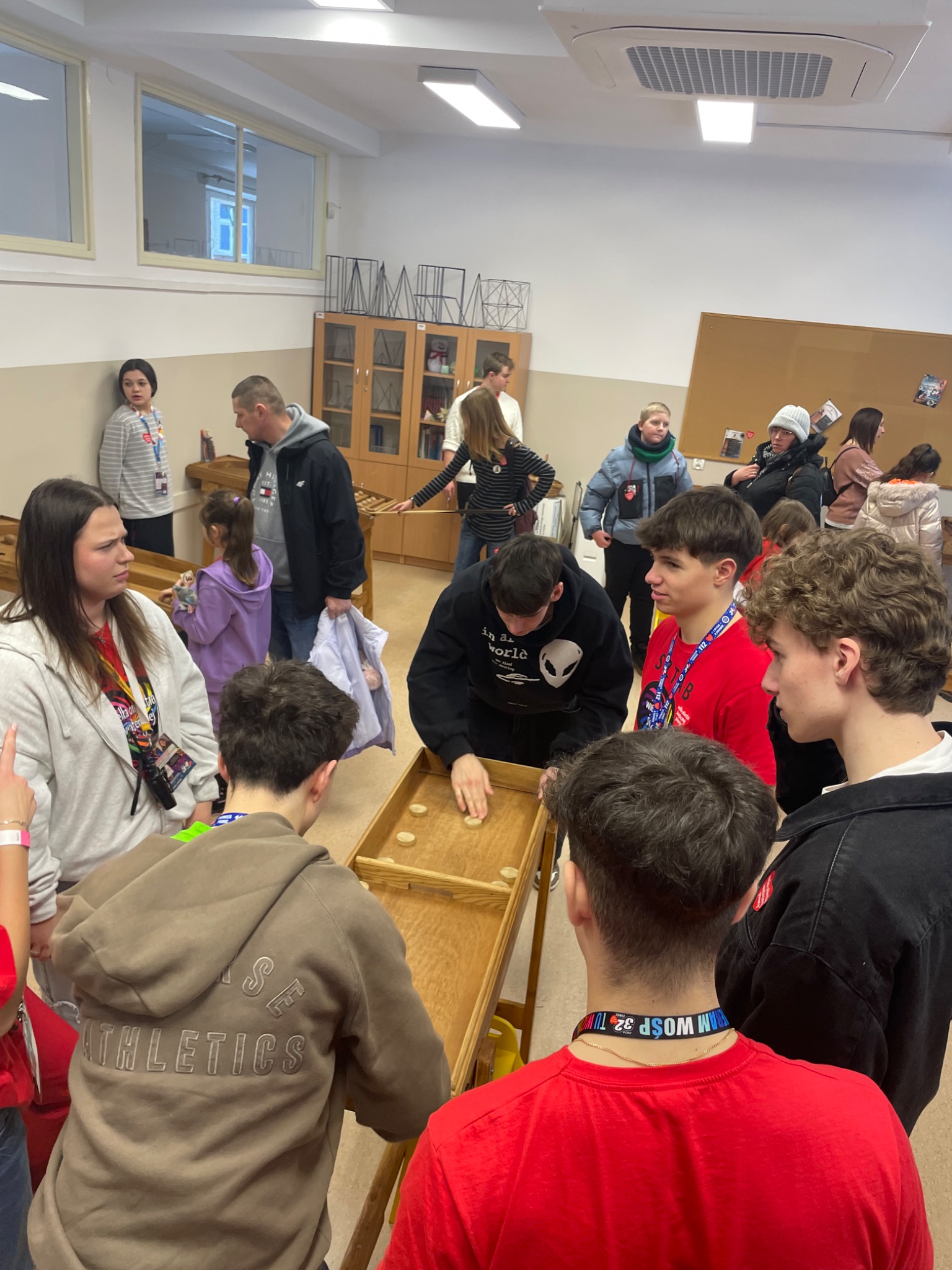 Gra w kapsle na drewnianym torze podczas wydarzenia. Uczestnicy w różnym wieku, w tle osoby obserwujące. Jasne oświetlenie, wnętrze sali.