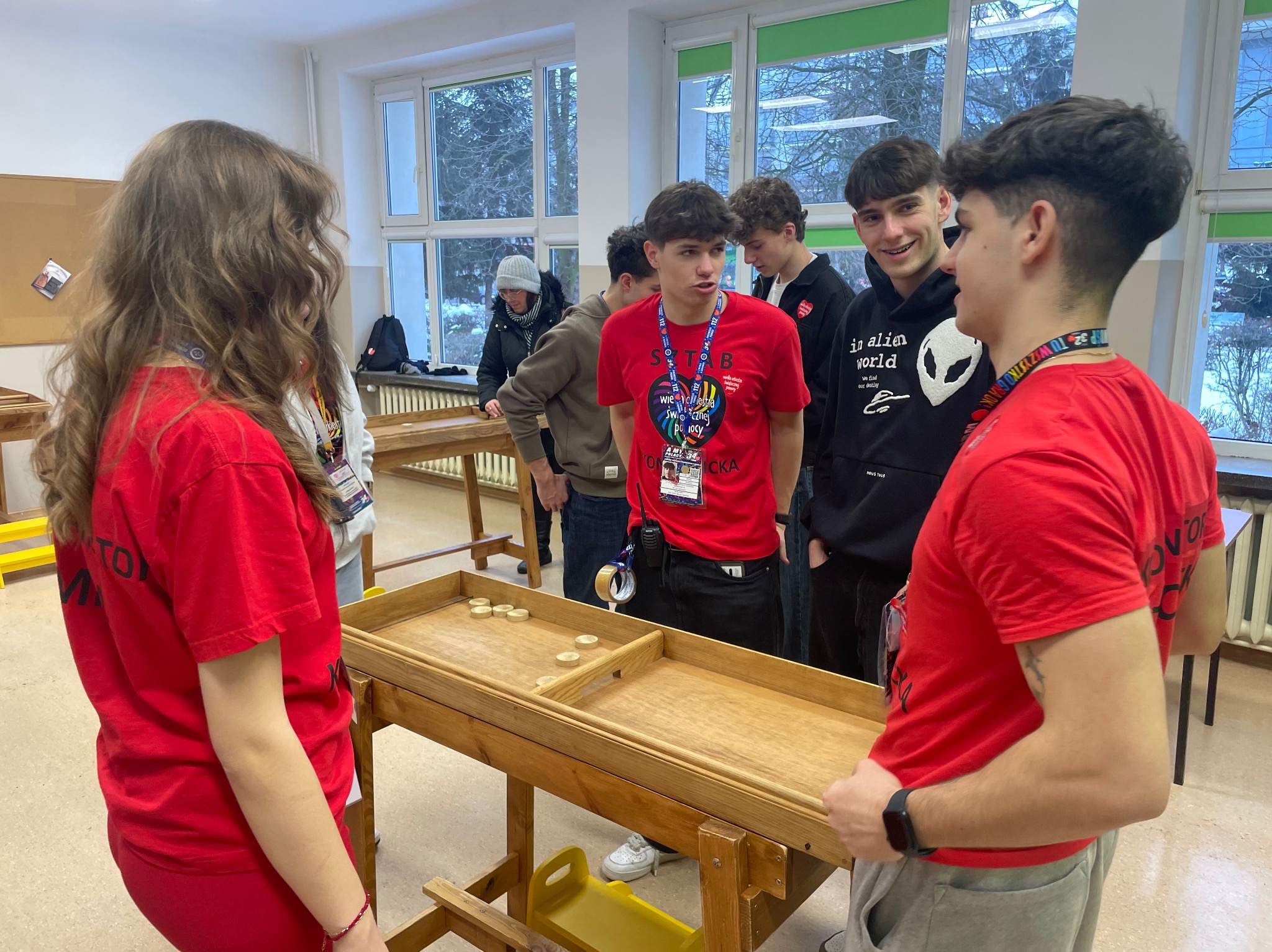 Młodzież w czerwonych koszulkach gra w grę z drewnianym stołem i krążkami w pomieszczeniu z oknami, prawdopodobnie podczas wydarzenia charytatywnego.