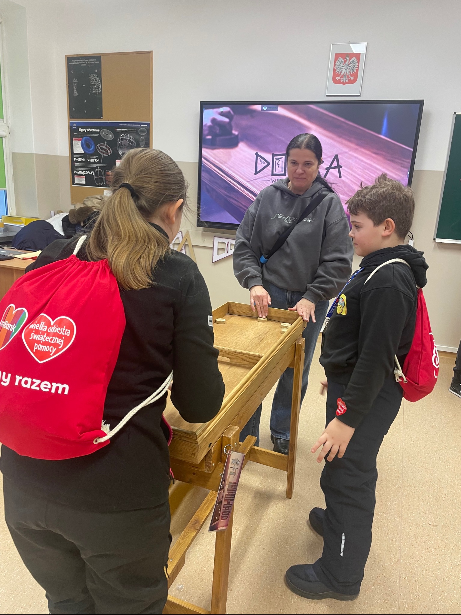 Uczestnicy WOŚP grają w grę z drewnianymi krążkami na stole, w tle ekran z logiem akcji. Dzieci z czerwonymi plecakami WOŚP, nauczycielka nadzoruje grę.