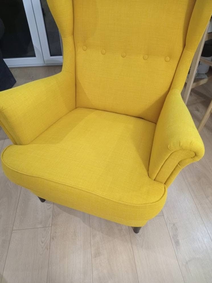 Żółty fotel typu uszak z tapicerką w zbliżeniu, widoczne detale wykończenia i faktura materiału. Ustawiony na jasnej podłodze, z fragmentem okna w tle.