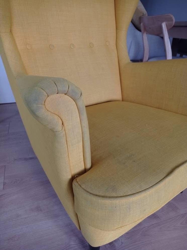 Żółty fotel tapicerowany z widocznymi plamami na podłokietniku i siedzisku, czekający na czyszczenie. W tle fragment innego mebla.