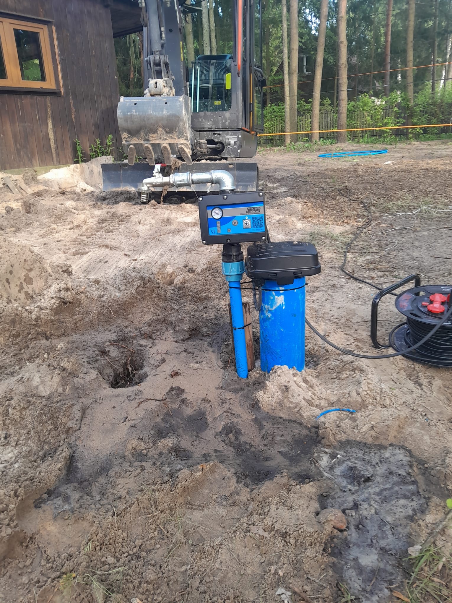 Instalacja gruntowa z niebieskimi rurami i czarną skrzynką sterującą, w tle koparka i drzewa. Widoczny etap prac przygotowawczych.
