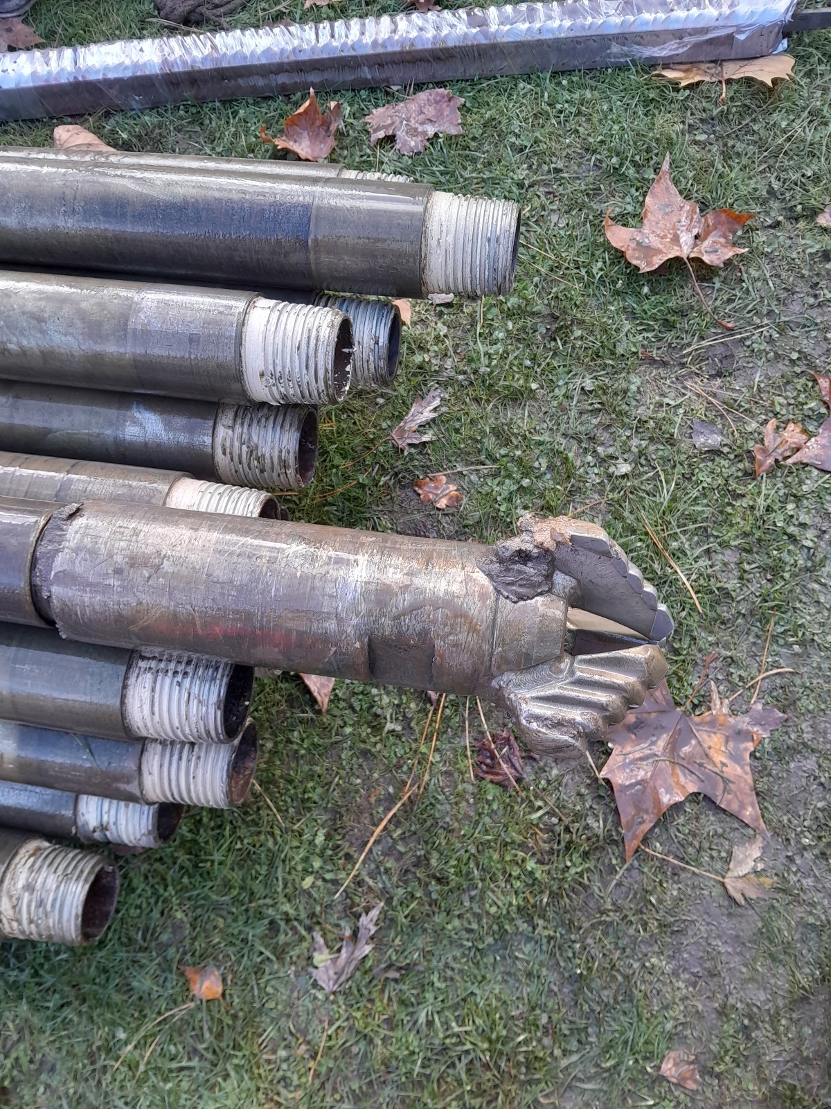 Wiertło do odwiertów geotermalnych leżące na trawie, widoczne gwintowane końcówki rur wiertniczych. Urządzenie do wykonywania odwiertów pod instalacje grzewcze.