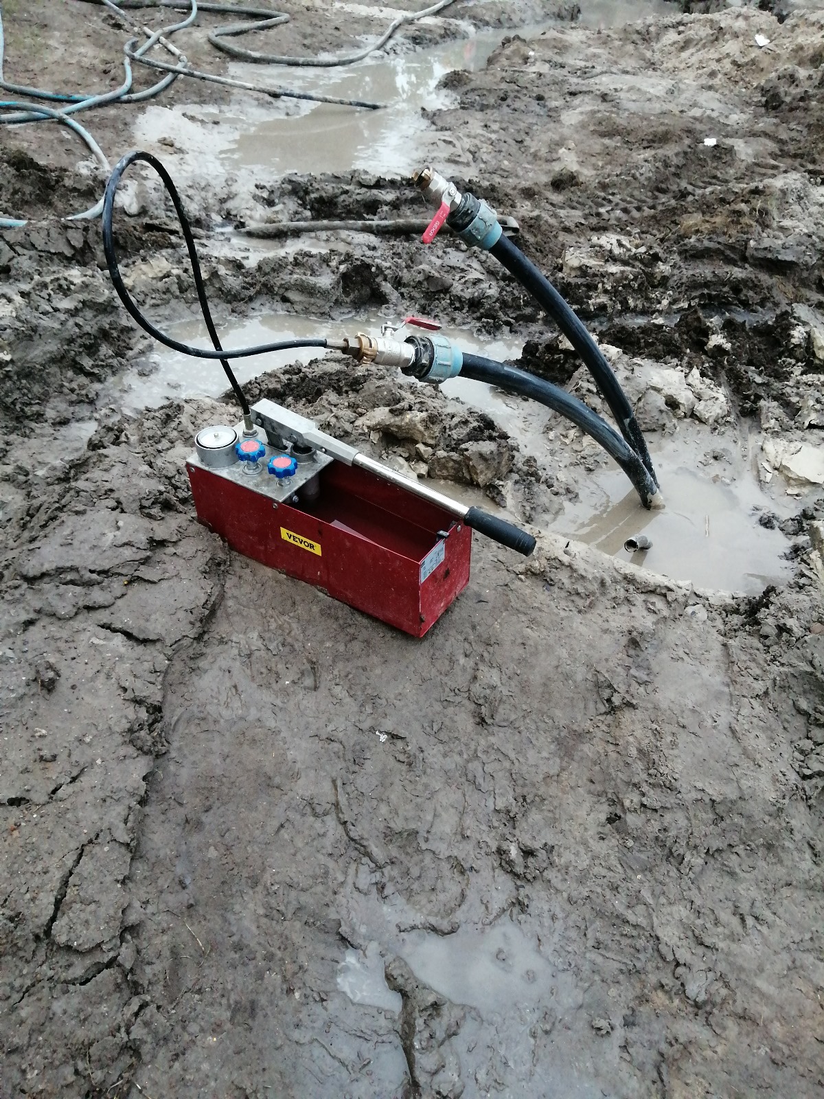 Czerwona pompa ciśnieniowa Vevor podłączona do rur w błotnistym terenie, prawdopodobnie podczas testu szczelności instalacji pompy ciepła.
