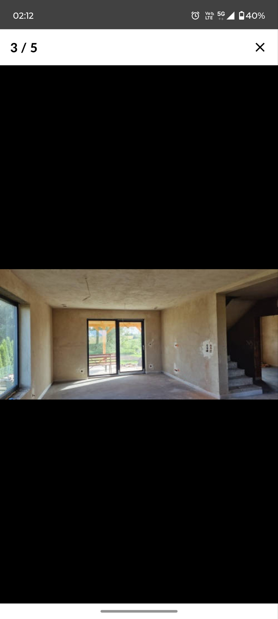 Surowe wnętrze domu w trakcie wykończenia: ściany po szpachlowaniu, widoczne instalacje elektryczne, schody w stanie surowym, duże okna z widokiem na zewnątrz.