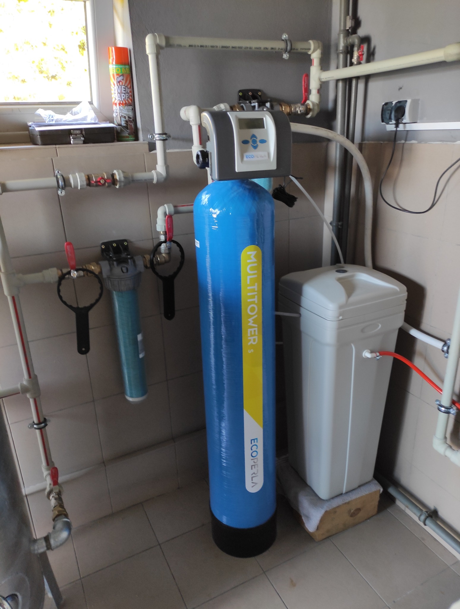 Zestaw filtrów do wody EcoPerla Multitower z podłączoną instalacją hydrauliczną w pomieszczeniu z szarą glazurą. Widoczne rury PCV z zaworami i przyłącza elektryczne.