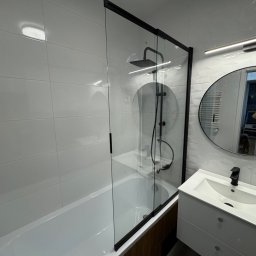 REN Bud - Nowoczesna łazienka po remoncie: białe płytki, czarna armatura, szklana kabina prysznicowa z deszczownicą i okrągłe lustro z oświetleniem LED. Widoczne odbicie osoby robiącej zdjęcie.