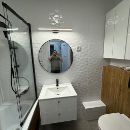 REN Bud - Nowoczesna łazienka z kabiną prysznicową typu walk-in, okrągłym lustrem i białymi szafkami. Na ścianach białe płytki 3D. Widoczne elementy armatury w kolorze czarnym.