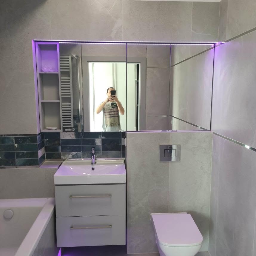 Szara łazienka po renowacji z podświetlanym lustrem szafkowym, umywalką z szafką i toaletą. Widoczne płytki imitujące marmur i element dekoracyjny z ciemnozielonej mozaiki.