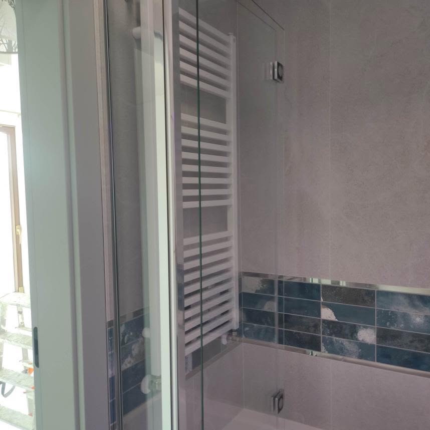 Nowoczesna łazienka z grzejnikiem drabinkowym za szklaną kabiną prysznicową. Widoczne płytki w odcieniach szarości i błękitu oraz fragment okna.