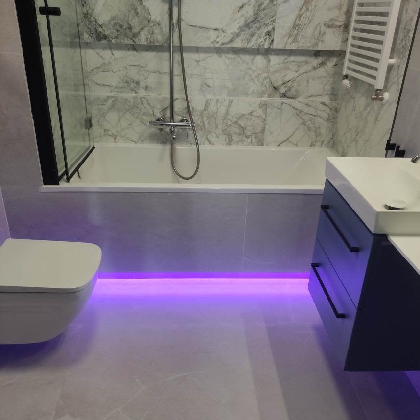 Nowoczesna łazienka z podświetleniem LED pod wanną i szafką umywalkową. Płytki imitujące marmur na ścianach. Kabina prysznicowa z czarną ramą.