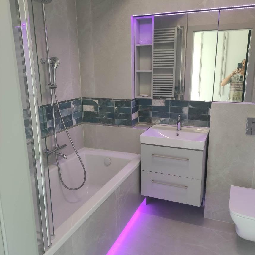 Wykończona łazienka w szarościach z akcentem niebieskich płytek i oświetleniem LED pod szafką umywalkową oraz lustrem. Widoczna wanna z prysznicem i toaleta.