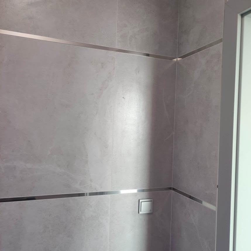 Ściana wykończona szarymi płytkami z marmurkowym wzorem, oddzielona poziomymi, metalowymi listwami. Widoczny włącznik światła i fragment futryny drzwi.