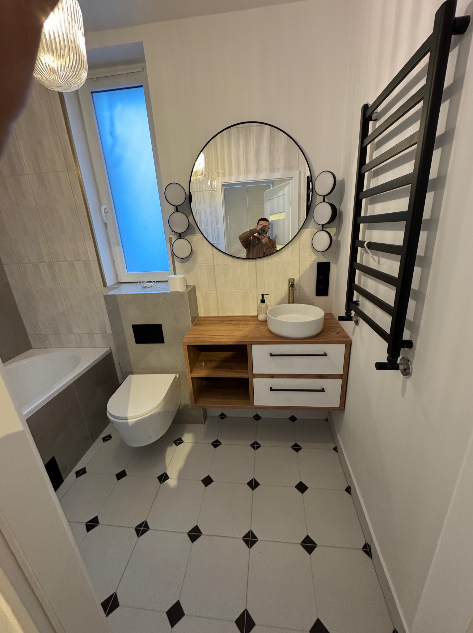 Nowoczesna łazienka z okrągłym lustrem, drewnianą szafką pod umywalką i czarnym grzejnikiem drabinkowym. Białe płytki podłogowe z czarnymi akcentami.