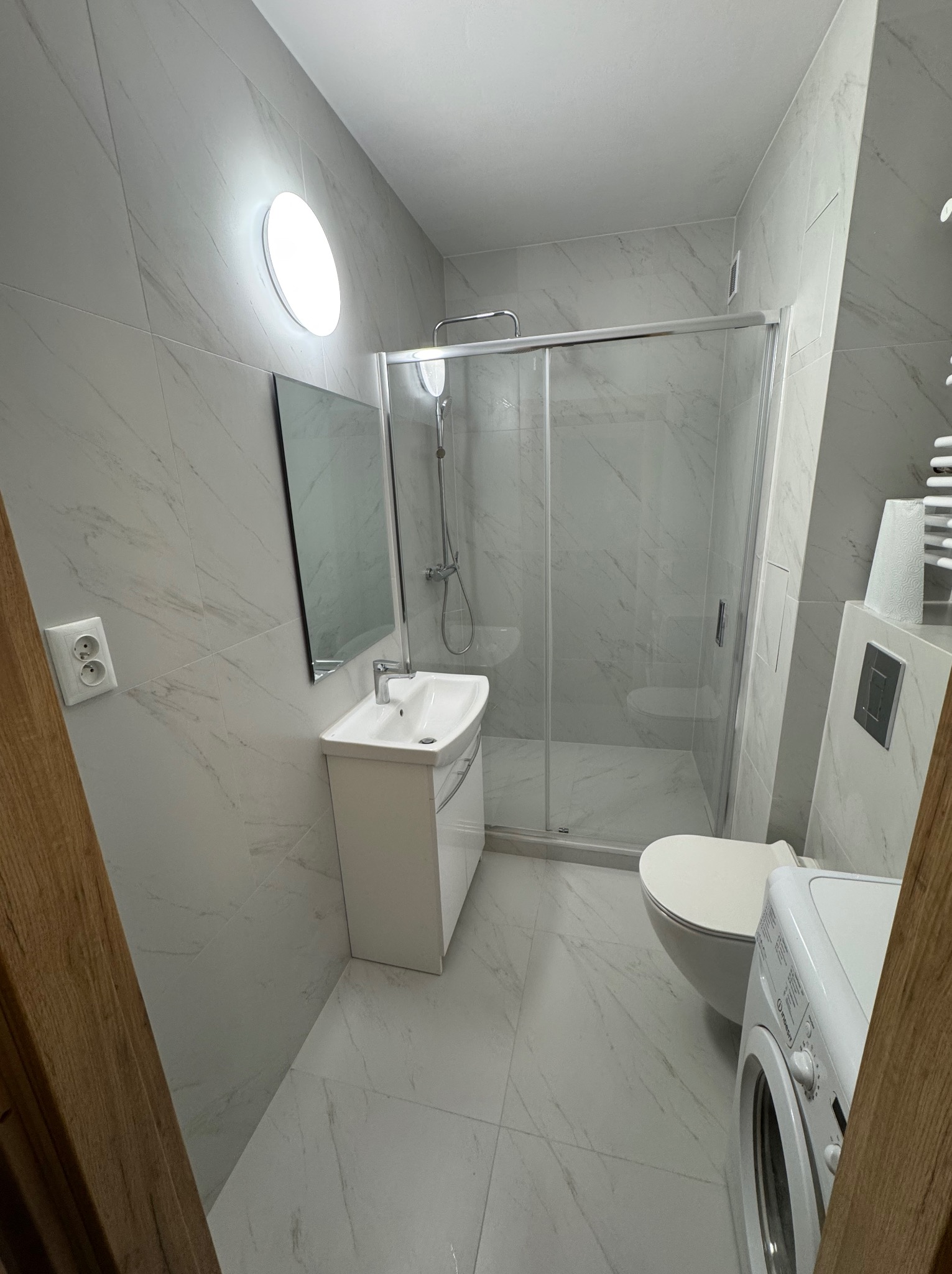 Jasna łazienka z kabiną prysznicową, umywalką z szafką i pralką. Ściany i podłoga wyłożone płytkami imitującymi marmur. Okrągłe oświetlenie sufitowe.