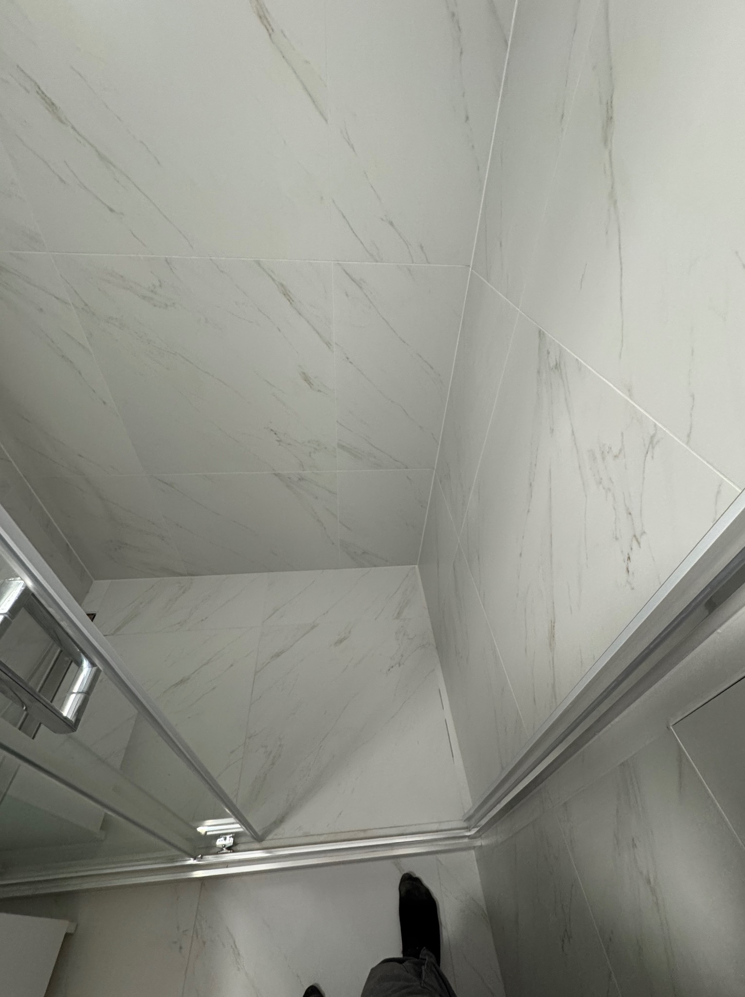 Wykończona kabina prysznicowa z białymi płytkami imitującymi marmur na ścianach i podłodze. Widoczny fragment osoby w dolnej części kadru.