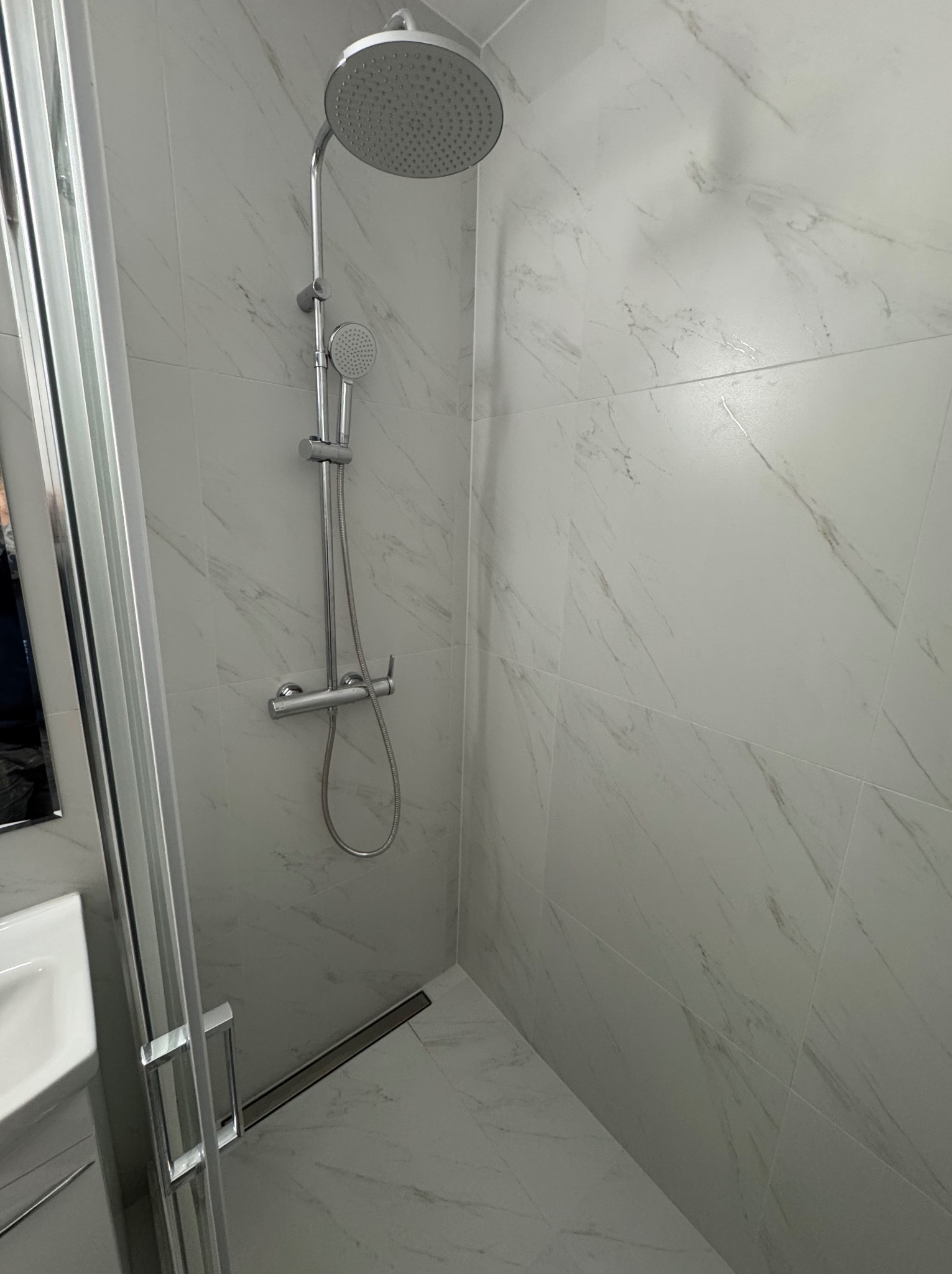 Wykończona kabina prysznicowa z deszczownicą i odpływem liniowym, wyłożona białymi płytkami imitującymi marmur. Widoczny fragment umywalki i szklane drzwi kabiny.