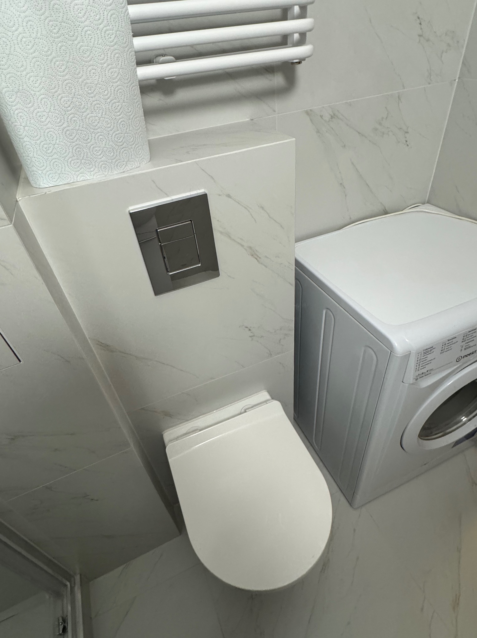 Nowoczesna łazienka z toaletą podwieszaną, pralką i grzejnikiem drabinkowym. Białe płytki imitujące marmur. Minimalistyczny design, czyste linie.