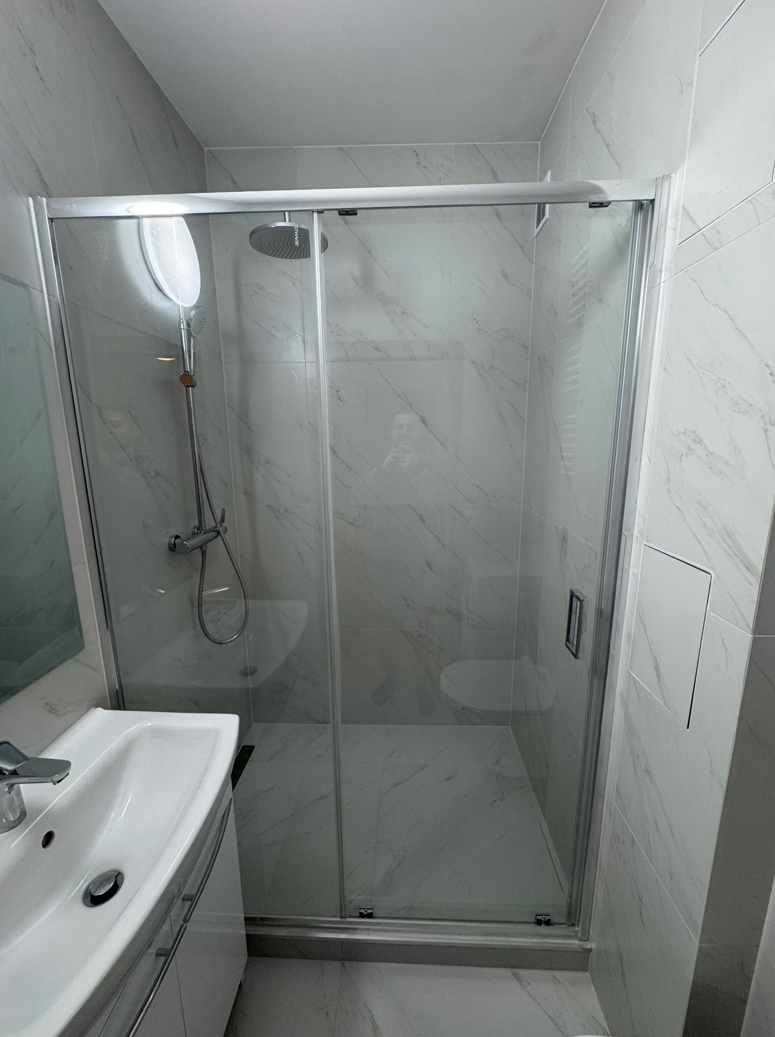 Nowoczesna kabina prysznicowa z przesuwnymi drzwiami, wykończona białymi płytkami imitującymi marmur, z widoczną umywalką i baterią w minimalistycznym stylu.