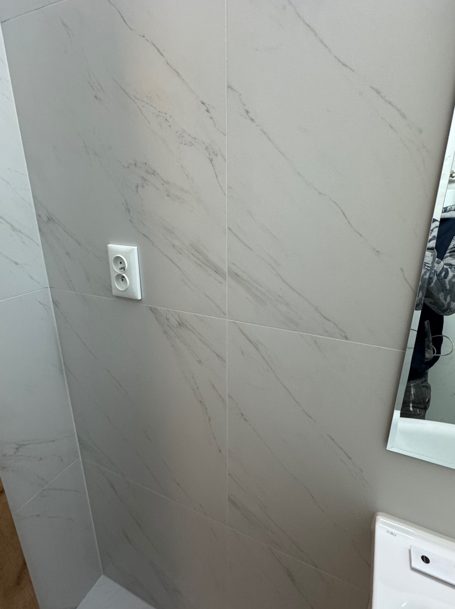 Ściana wyłożona jasnymi, marmurkowymi płytkami w łazience. Widoczne gniazdko elektryczne i fragment lustra z odbiciem osoby. Minimalistyczny design.