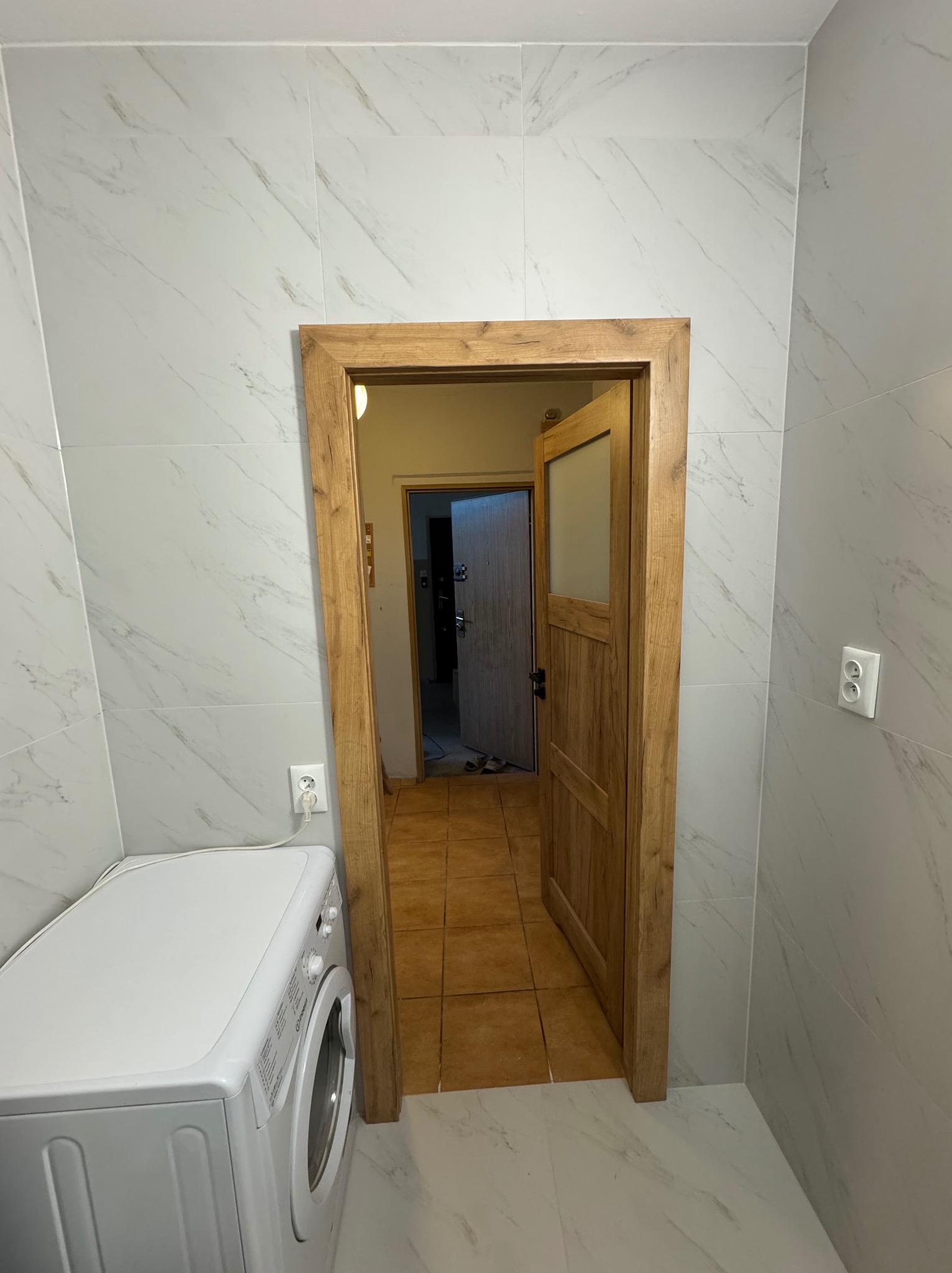 Wykończona łazienka z marmurowymi płytkami, drewnianą ościeżnicą i widoczną pralką. W tle otwarte drzwi do kolejnego pomieszczenia z brązową podłogą.