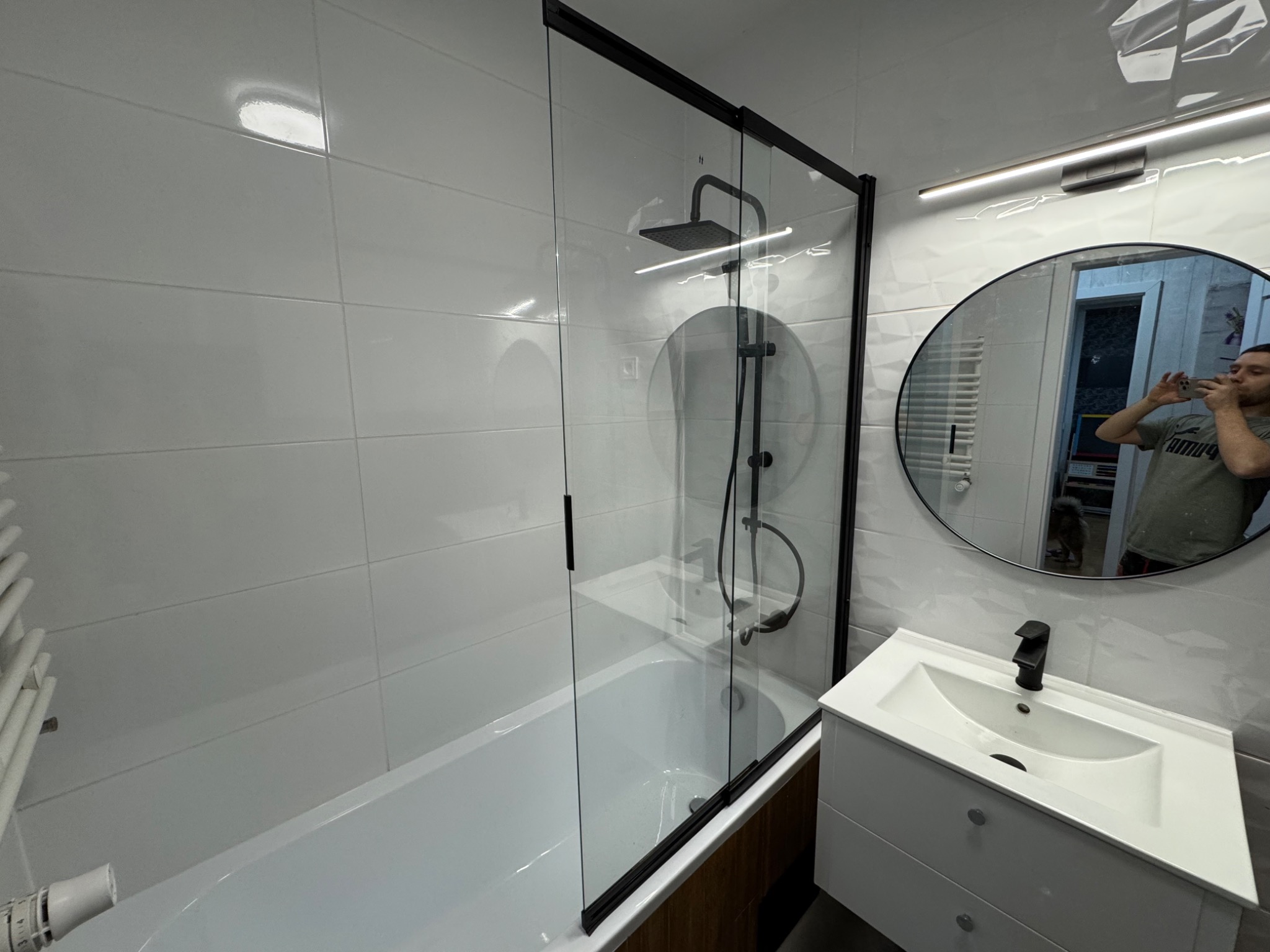 Nowoczesna łazienka po remoncie: białe płytki, czarna armatura, szklana kabina prysznicowa z deszczownicą i okrągłe lustro z oświetleniem LED. Widoczne odbicie osoby robiącej zdjęcie.
