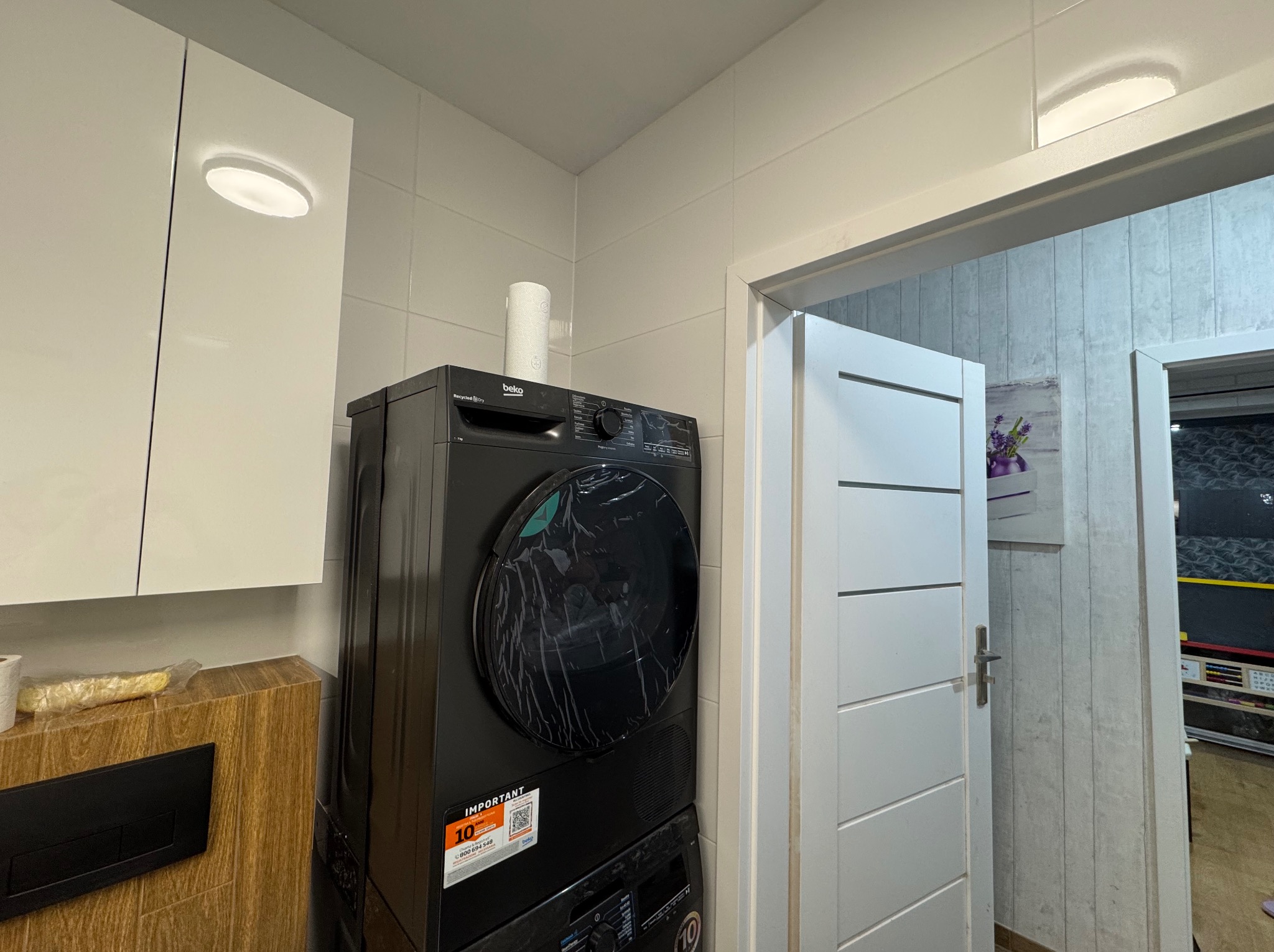 Nowoczesna łazienka z pralko-suszarką Beko w czarnej obudowie, białe płytki i szafki, widok na otwarte drzwi do innego pomieszczenia.