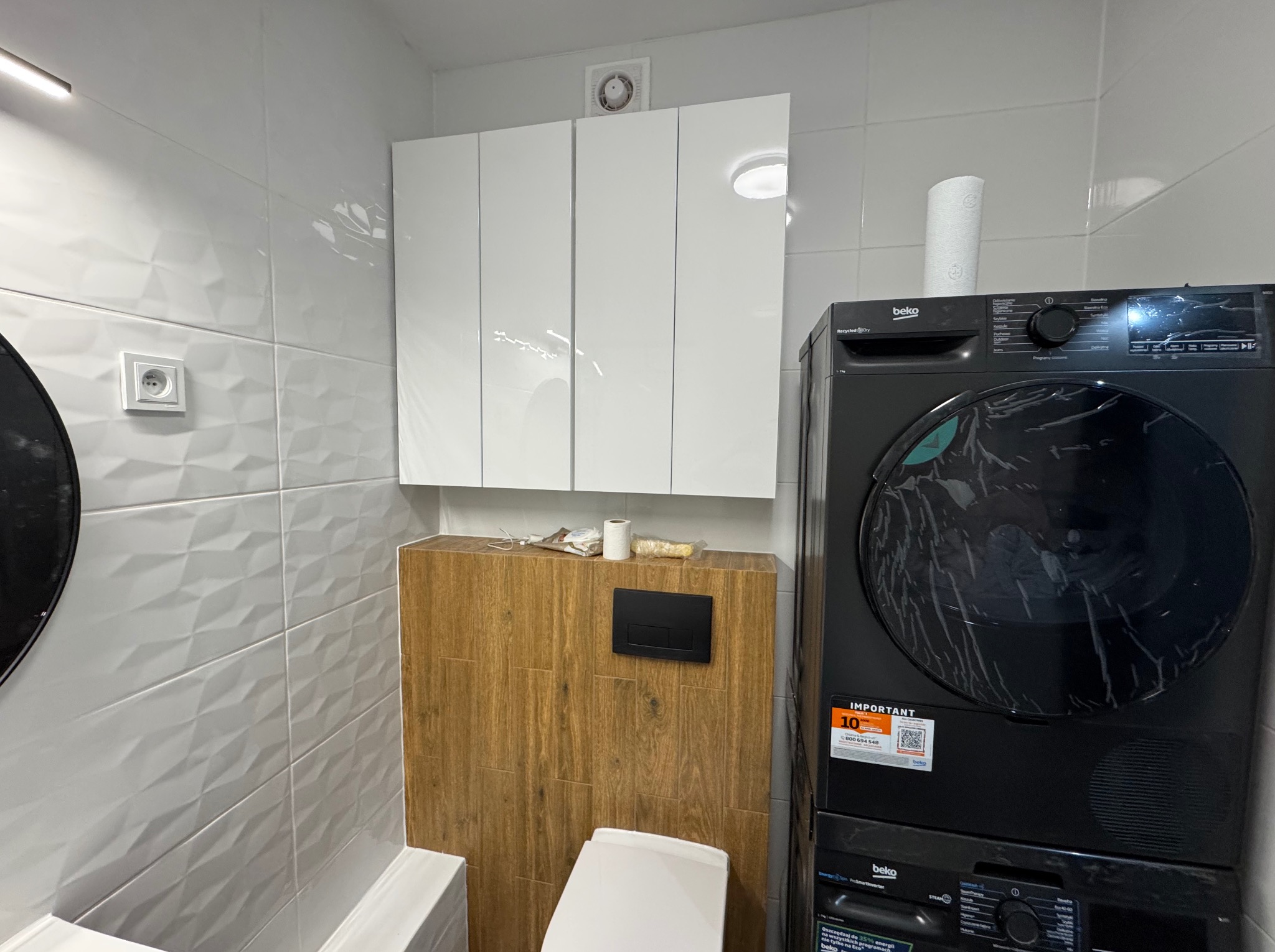 Nowoczesna łazienka po remoncie: białe, trójwymiarowe płytki, zabudowa WC z drewnopodobnymi panelami, białe szafki wiszące i pralka z suszarką Beko.