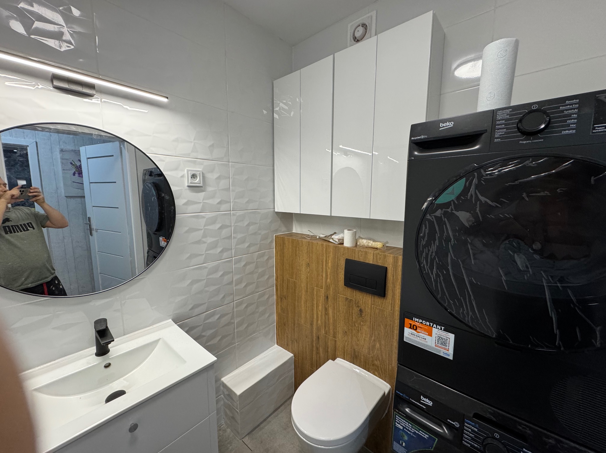 Nowoczesna łazienka po remoncie: białe płytki 3D, szafki wiszące, umywalka z lustrem, toaleta i pralko-suszarka Beko w czarnym kolorze. Widoczne detale wykończenia.