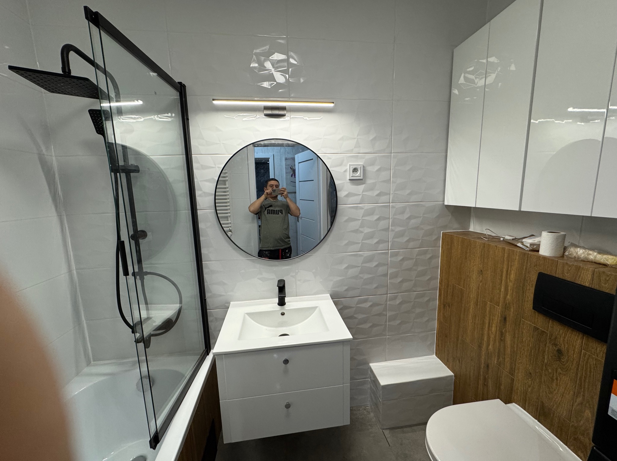 Nowoczesna łazienka z kabiną prysznicową typu walk-in, okrągłym lustrem i białymi szafkami. Na ścianach białe płytki 3D. Widoczne elementy armatury w kolorze czarnym.