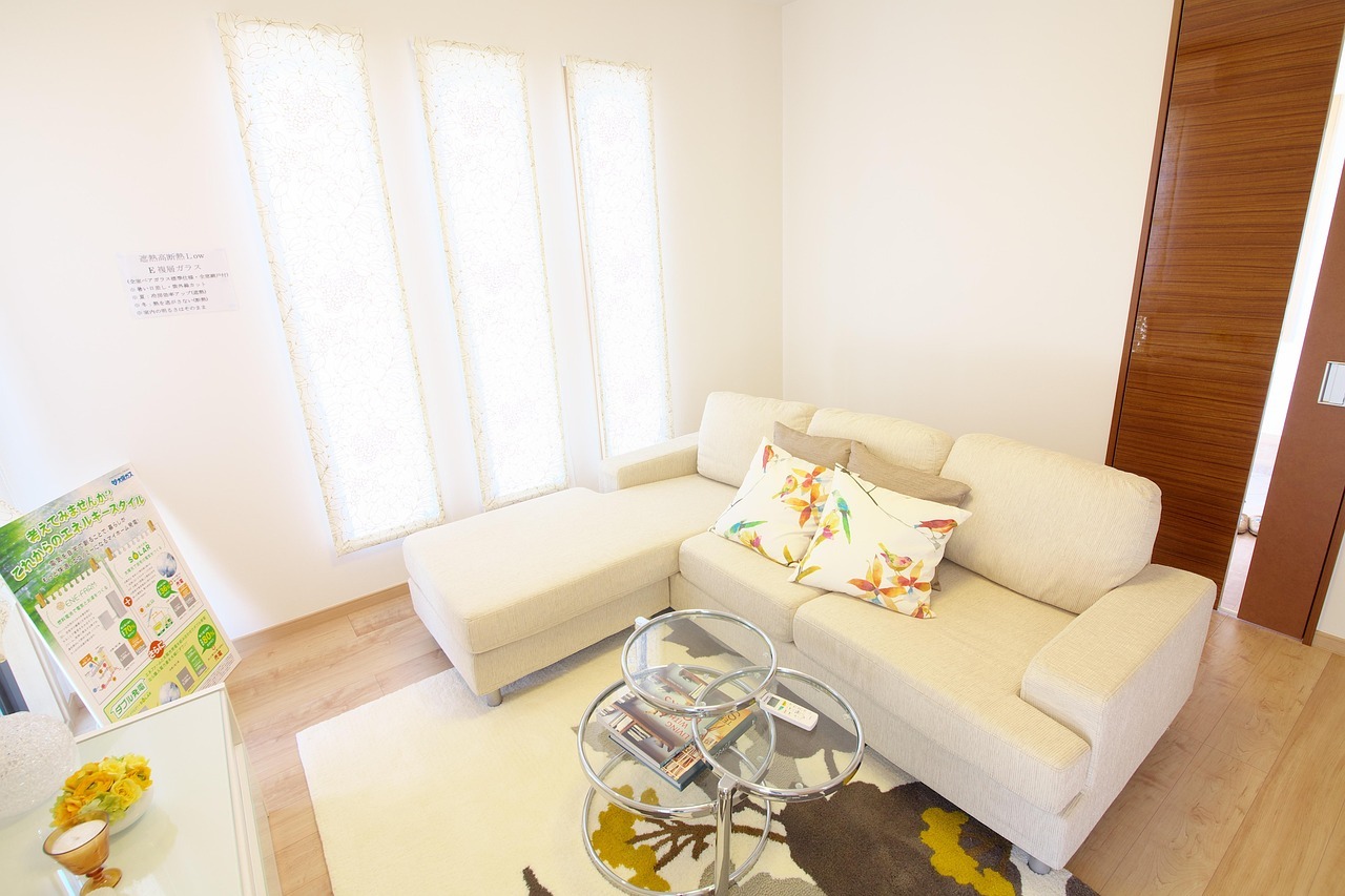 Jasne wnętrze: narożna sofa z poduszkami w kwiaty, stolik kawowy z trzema szklanymi blatami, wysokie okna z koronkowymi firanami, drewniane drzwi.