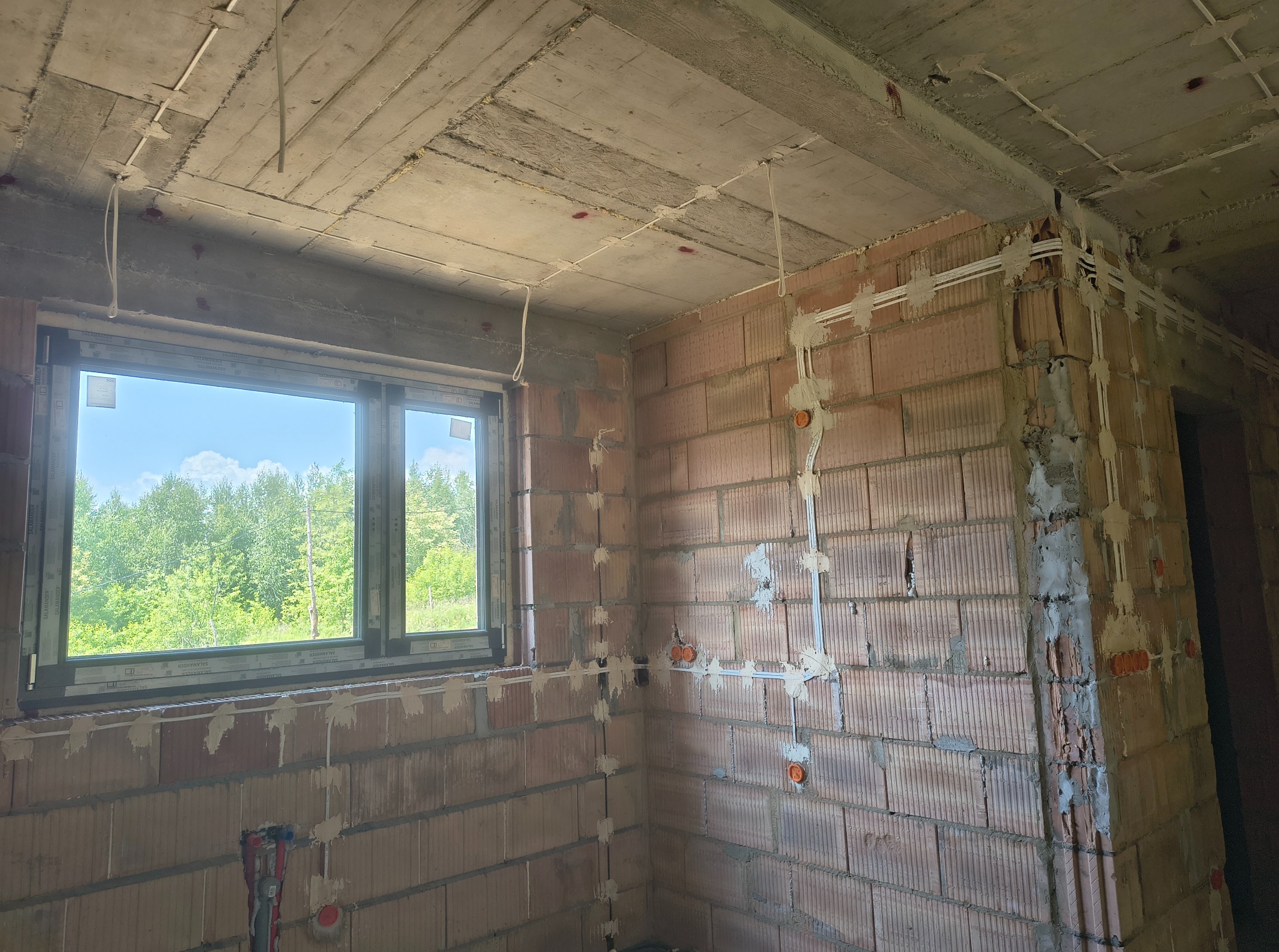 Surowy stan: instalacja elektryczna w budynku z pustaków, kable prowadzone po ścianach i suficie, widok na zieleń za oknem.