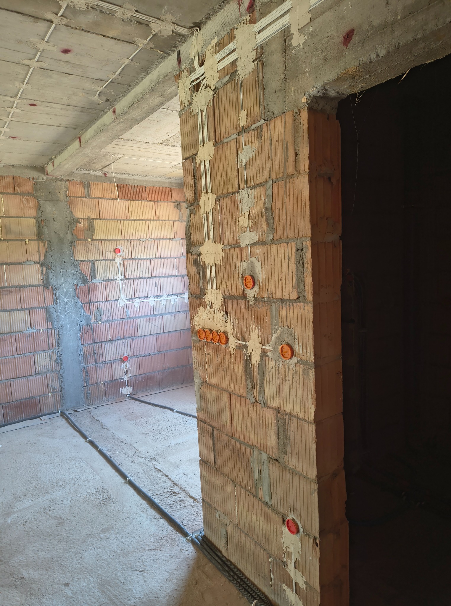 Surowy stan: instalacja elektryczna w bruzdach, pomarańczowe puszki w ścianie z cegły dziurawki, rury na podłodze i pod sufitem. Widok na wnętrze budynku w trakcie budowy.