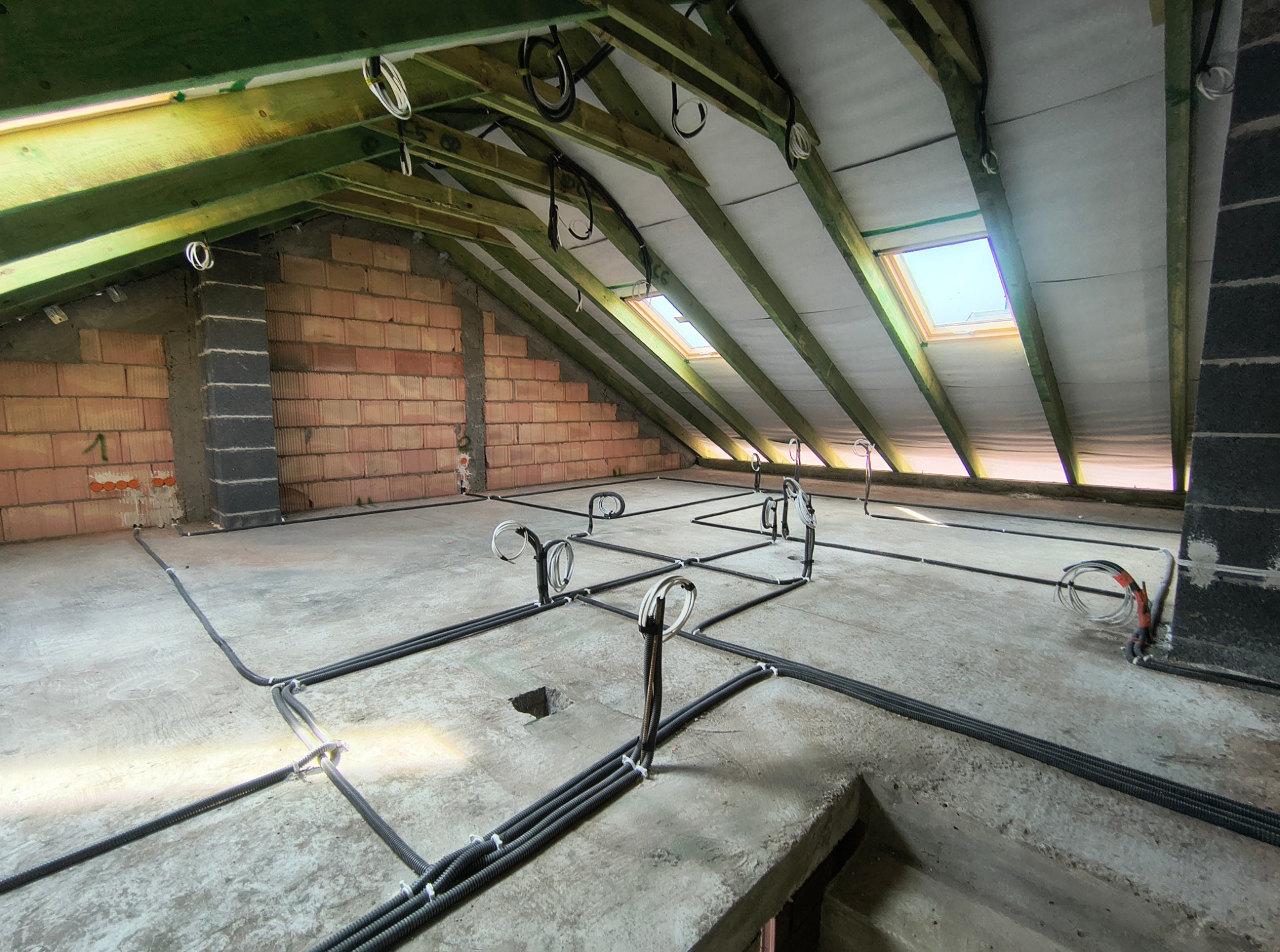 Rozprowadzone kable w peszlach na poddaszu, widoczne okna dachowe i ceglana ściana. Przygotowanie do instalacji elektrycznej w nowym domu.