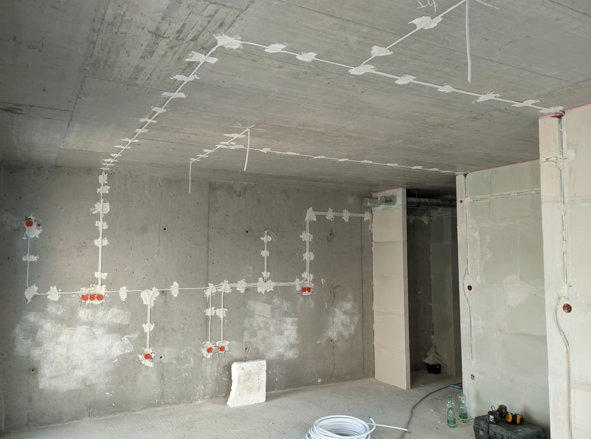 Surowy beton, widoczne białe kable elektryczne przytwierdzone do ścian i sufitu. Pomarańczowe puszki elektryczne w ścianach. W tle narzędzia i butelki.