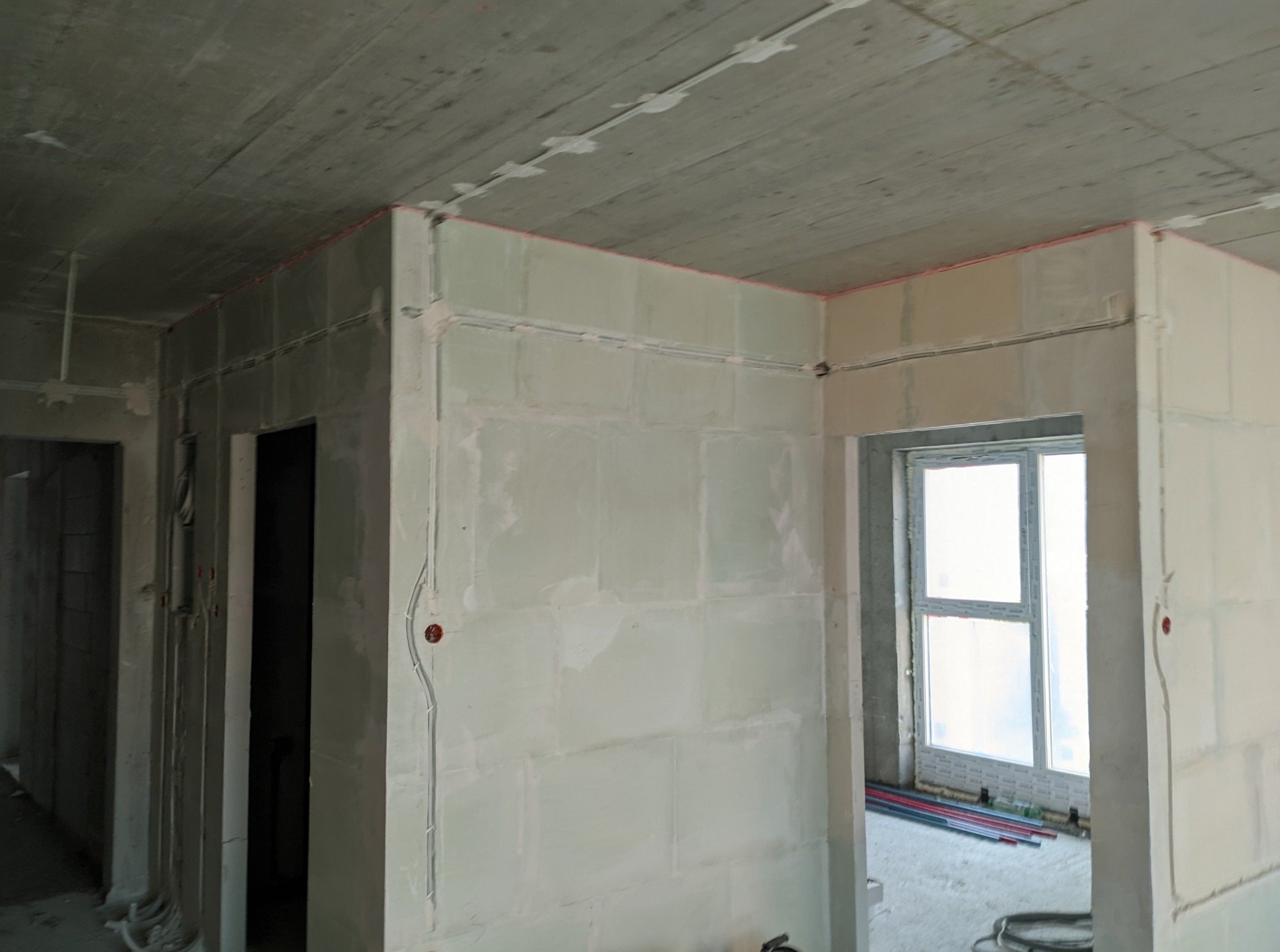 Wnętrze w stanie surowym: widoczne przewody elektryczne zamontowane na ścianach i suficie, przygotowane do dalszych prac wykończeniowych. Szare ściany, okno w tle.