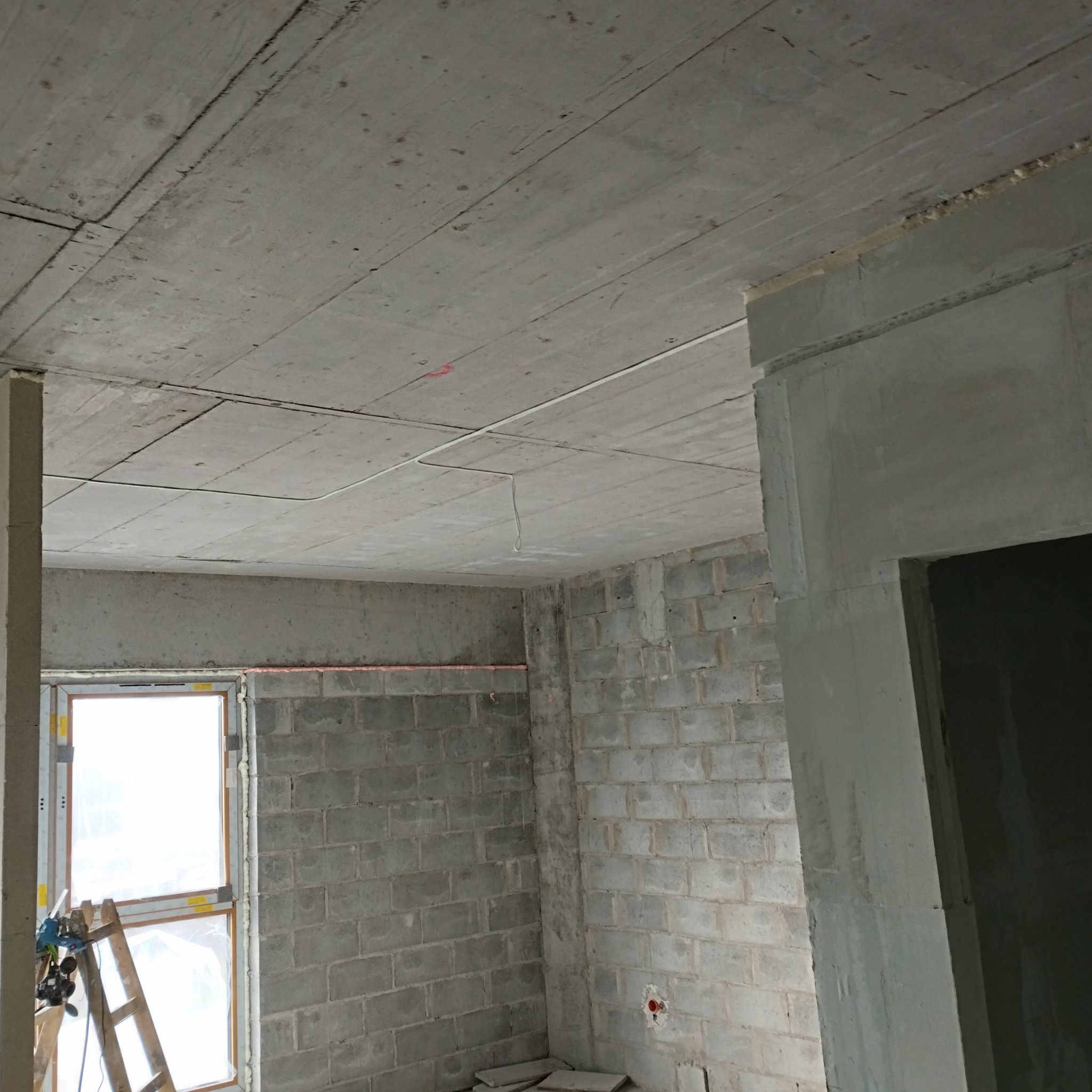 Surowy stan wnętrza: ściany z bloczków, okno z pianką montażową, drabina oparta, widoczne przewody pod sufitem, przygotowane pod instalacje.