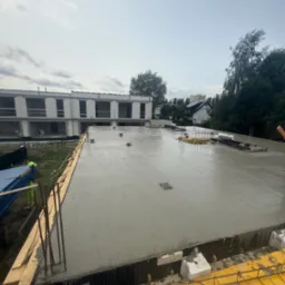 Świeżo wylany beton na stropie budynku w trakcie budowy, widoczne szalunki i zbrojenia. W tle budynek w stanie surowym otwartym i zieleń.