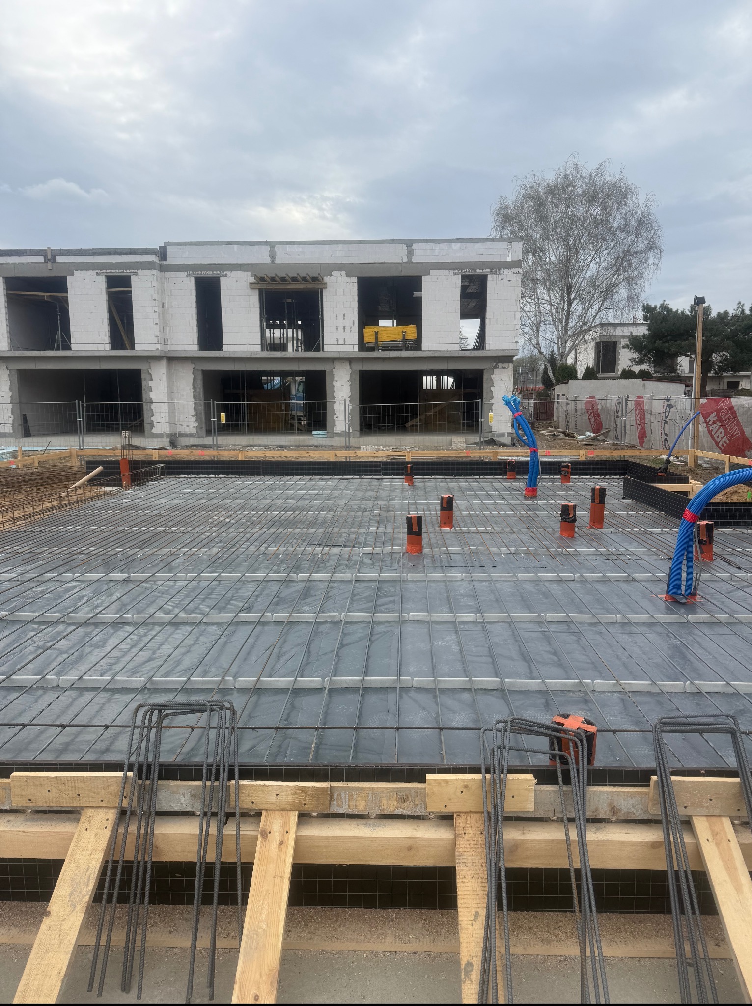 Zbrojenie płyty fundamentowej pod budynek z betonu komórkowego. Widoczne wystające rury instalacyjne i drewniane szalunki. Pochmurne niebo w tle.