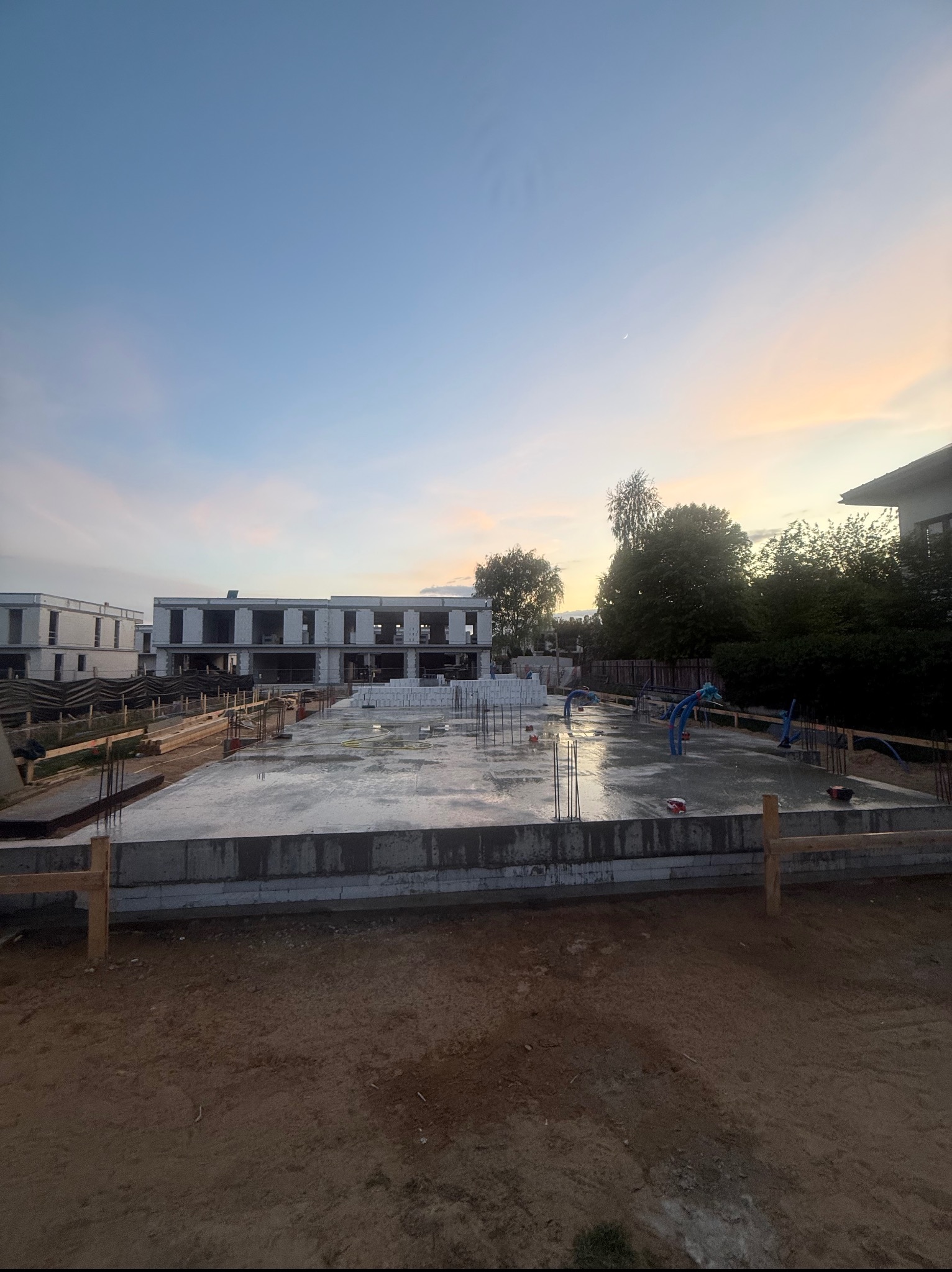 Wylana, mokra płyta fundamentowa pod budowę domu jednorodzinnego z wystającymi prętami zbrojeniowymi i instalacjami. W tle budynki w trakcie budowy.