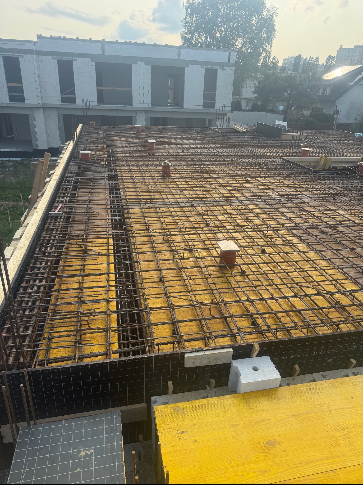 Zbrojenie stropu budynku w trakcie budowy. Widoczne żółte szalunki, siatka zbrojeniowa i fragment ściany z pustaków. W tle budynki mieszkalne i drzewa.