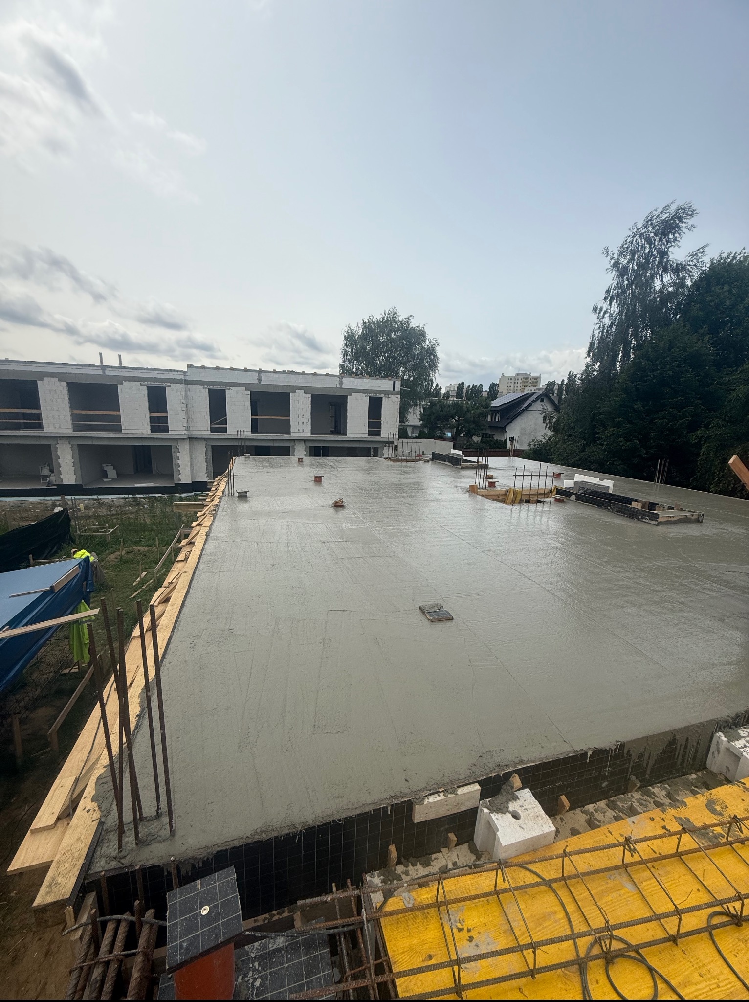 Świeżo wylany beton na stropie budynku w trakcie budowy, widoczne szalunki i zbrojenia. W tle budynek w stanie surowym otwartym i zieleń.