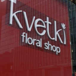 AdverHome Aksana Halaveika - Elegancki szyld 3D w kolorze białym na czerwonym tle, z napisem 'Kvetki floral shop', umieszczony na szklanej fasadzie budynku, odbijający miejski krajobraz.
