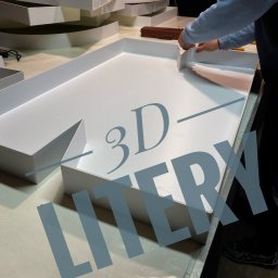 AdverHome Aksana Halaveika - Ręczne składanie białych liter 3D na stole warsztatowym, widoczne fragmenty innych liter i narzędzia. Oświetlenie studyjne, praca w toku.