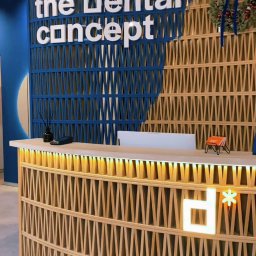 AdverHome Aksana Halaveika - Recepcja gabinetu stomatologicznego z drewnianą ażurową ścianą w kolorze niebieskim i beżowym, podświetlonym logo i świątecznym wieńcem.